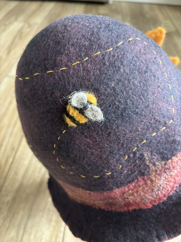 Close up of a bee felted onto a cloche hat with a dashed line of embroidery marking the bee's path around the hat