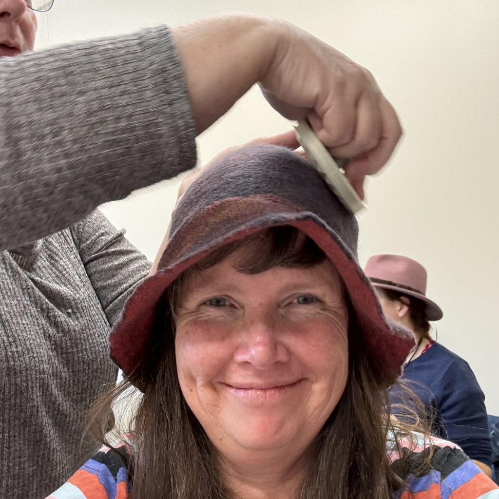 A girl smiles into the camera with a felted cloche hat on her head while the instructor rubs it with a small plastic disk