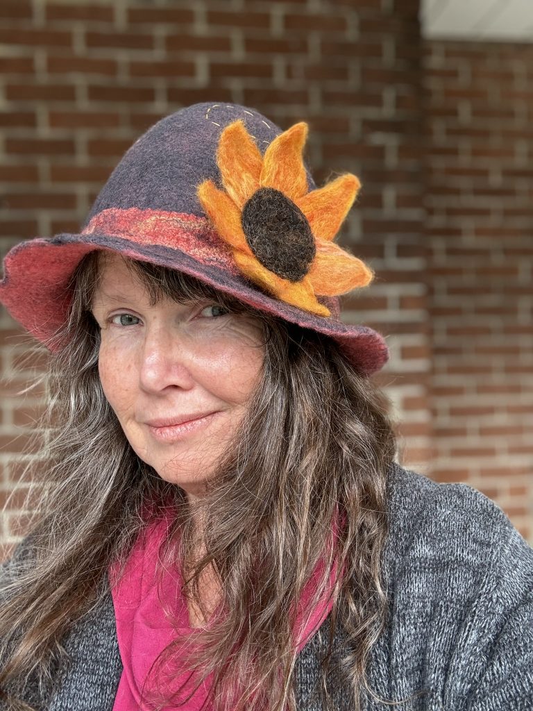 A middle-aged woman with long brown hair smirks slightly while wearing a grey cloche hat with a big sunflower on the brim