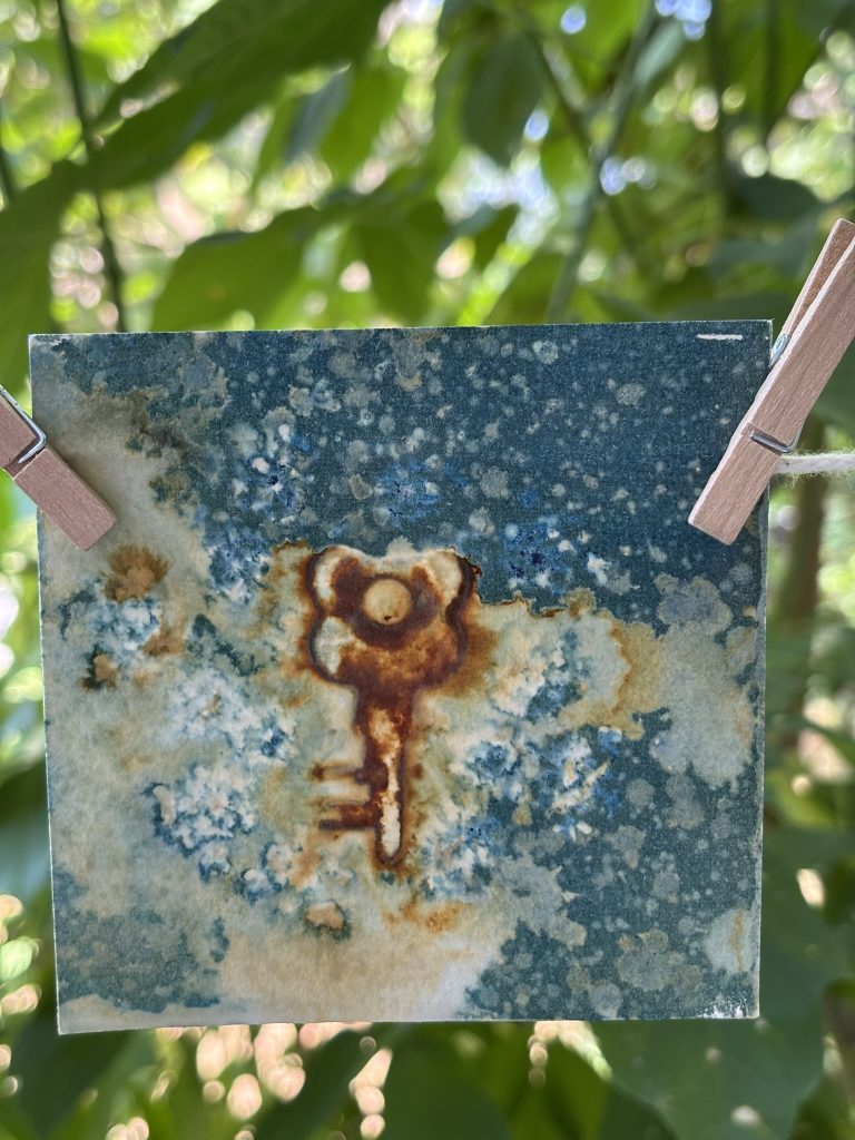 A wet cyanotype hangs to dry on a line, with swirls of rusty brown and blue in organic shapes around the outlines of vintage keys