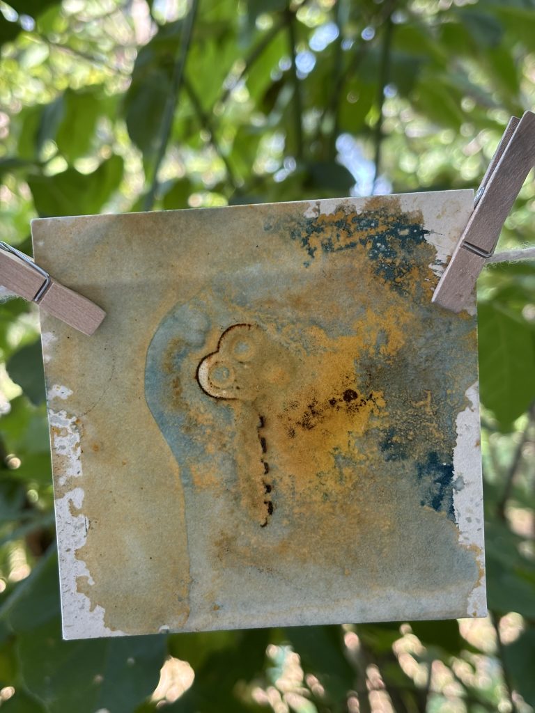 A wet cyanotype hangs to dry on a line, with swirls of rusty brown and blue in organic shapes around the outlines of vintage keys