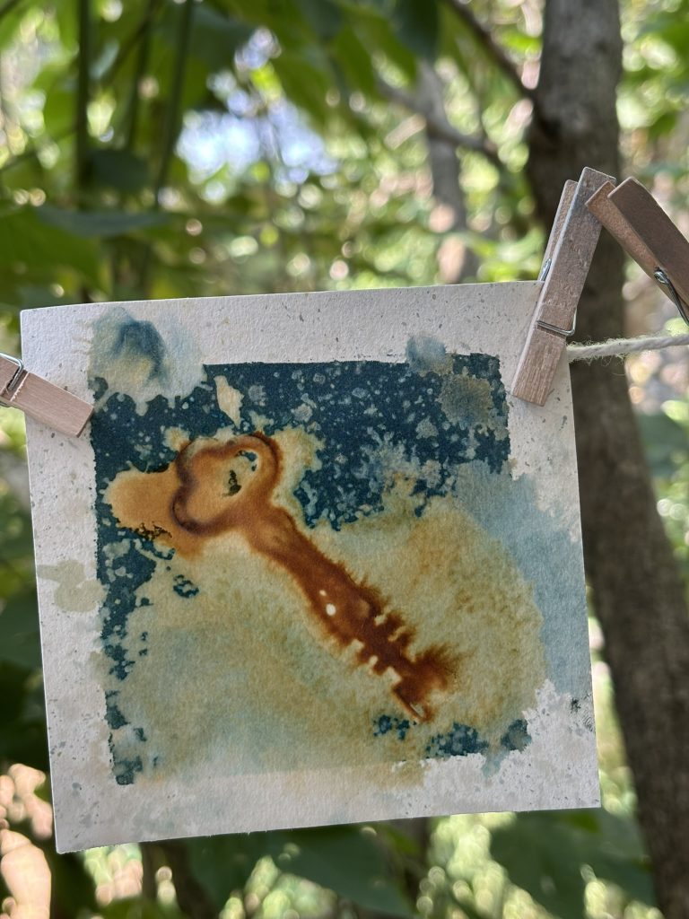 A wet cyanotype hangs to dry on a line, with swirls of rusty brown and blue in organic shapes around the outlines of vintage keys