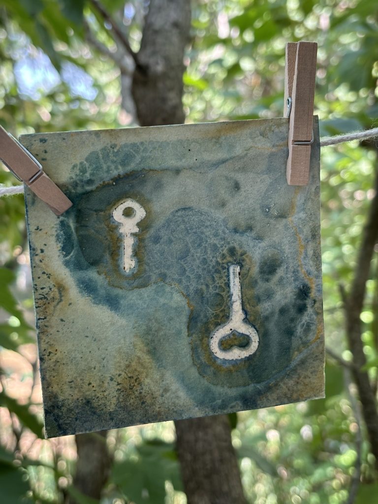 A wet cyanotype hangs to dry on a line, with swirls of rusty brown and blue in organic shapes around the outlines of vintage keys