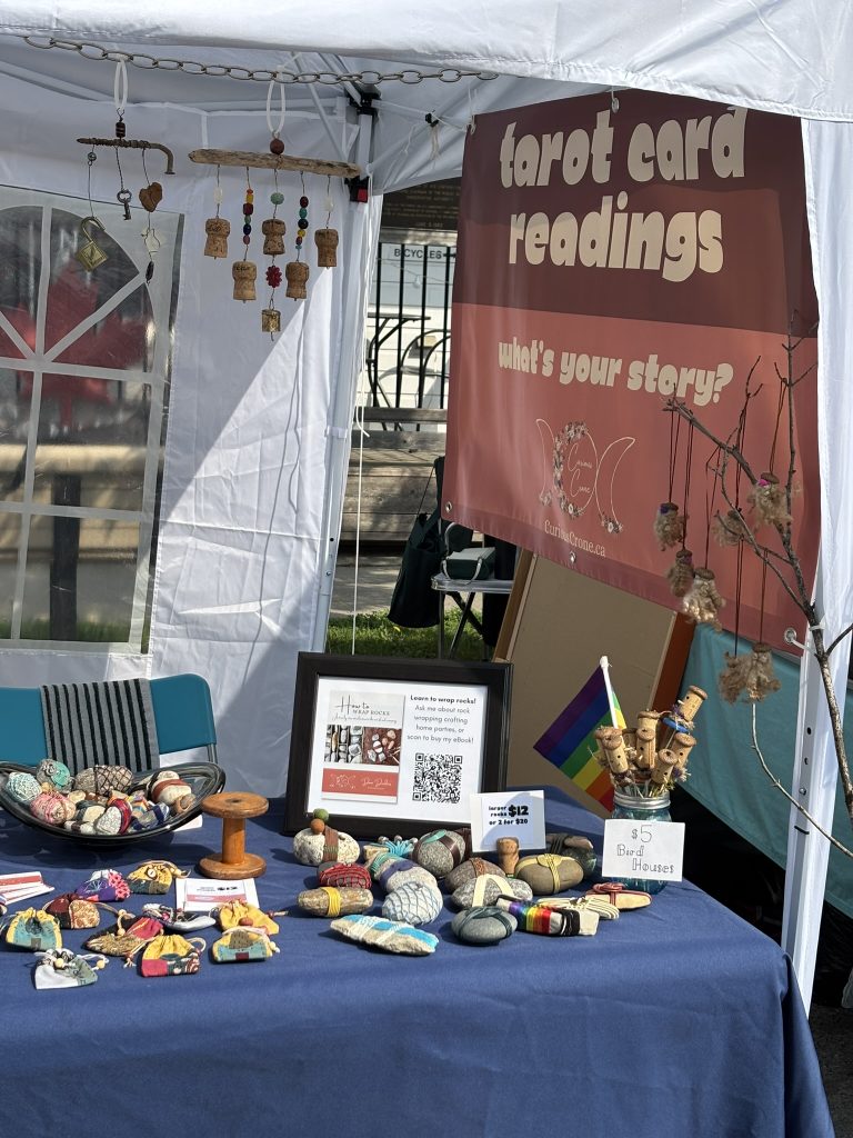 A table at a craft show set up with folk art crafts and a sign that says "Tarot readings - what's your story?"